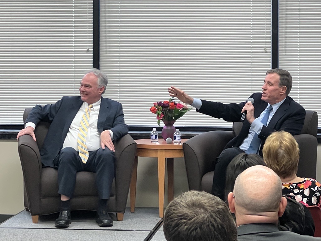 Senator Mark Warner talking to a room full of people with Senator Tim Kaine seated nearby
